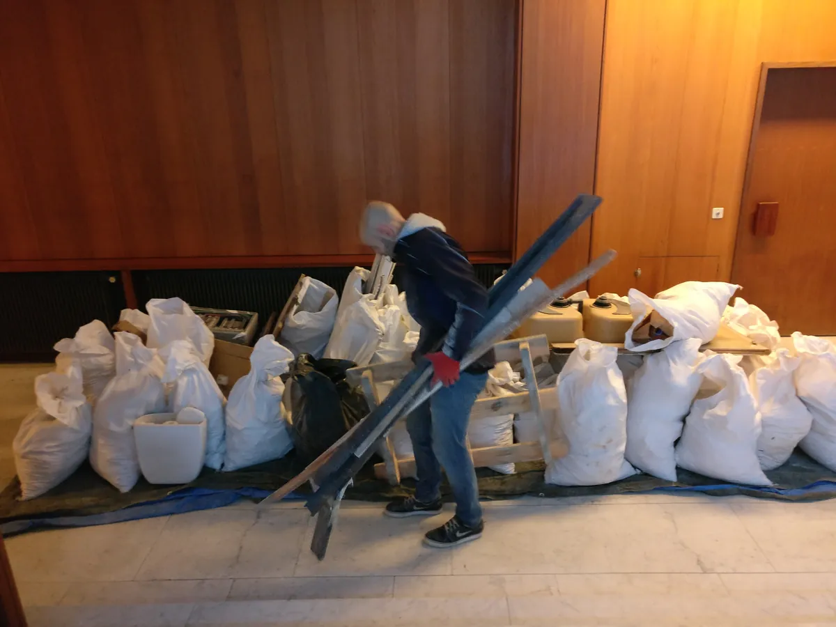 Homme portant des chutes de bois dans une salle lambrissée, sacs blancs, WC, sac noir et bidons alignés le long du mur