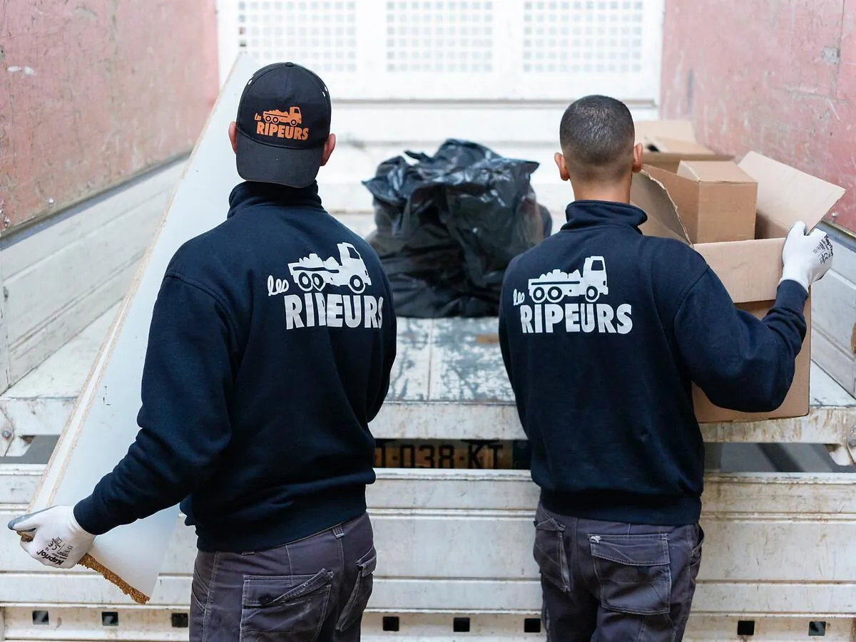Deux ripeurs de dos, veste Les Ripeurs, chargeant des cartons sur le plateau d'un camion depuis une cour