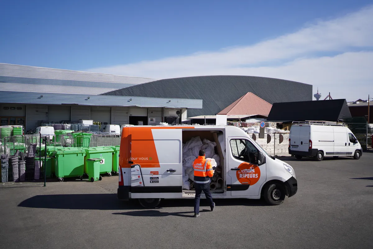 Fourgon Les Ripeurs porte ouverte, chargé de sacs blancs, sur une plateforme de valorisation avec bacs verts