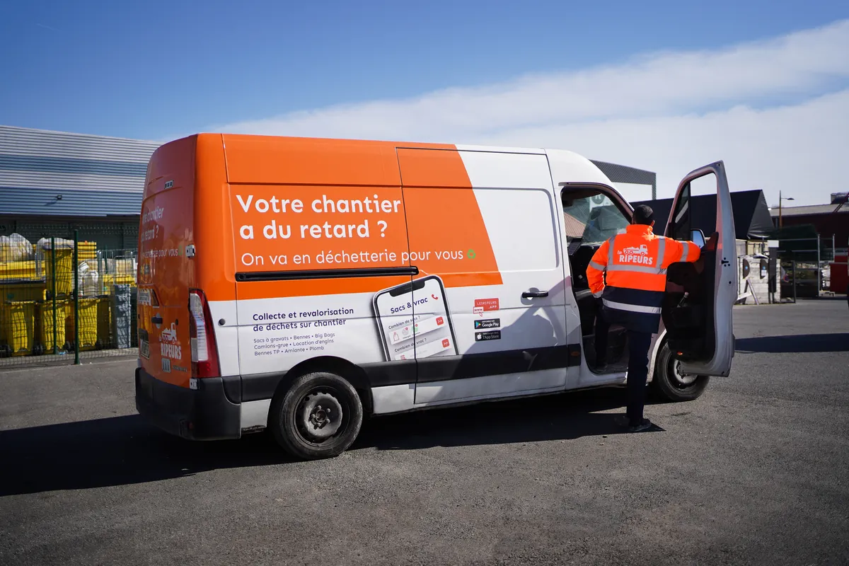Fourgon Les Ripeurs orange et blanc portant le message « Votre chantier a du retard ? On va en déchetterie pour vous », ripeur montant à bord