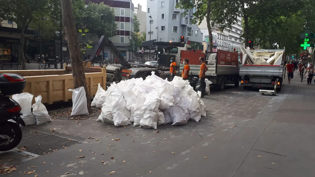 Gros tas de sacs blancs sur un trottoir en centre-ville, benne jaune et deux camions Les Ripeurs, équipe en orange au travail