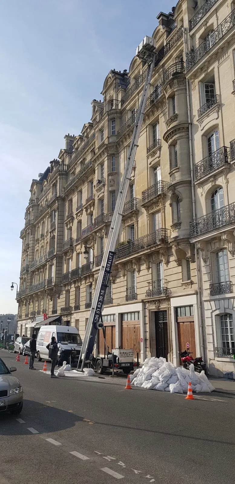Monte-charge Giralift déployé sur une façade haussmannienne de quai parisien, tas de sacs blancs et cônes au pied, fourgon Les Ripeurs.
