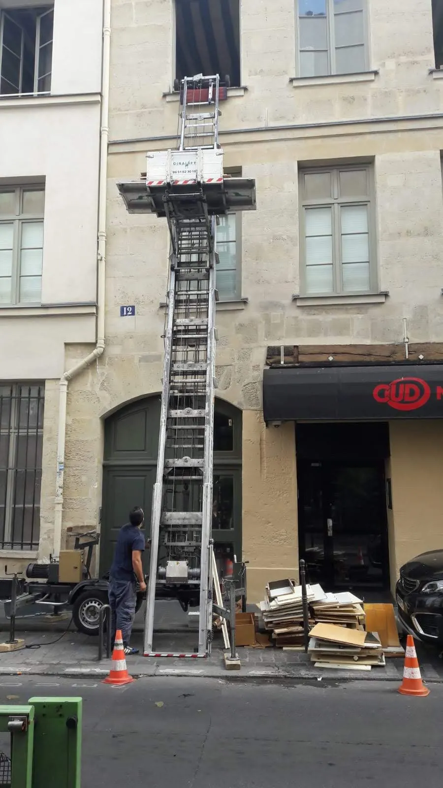 Monte-charge sur remorque dressé contre un immeuble ancien dans une rue d'immeubles anciens, panneaux de mobilier empilés au trottoir et cônes de signalisation