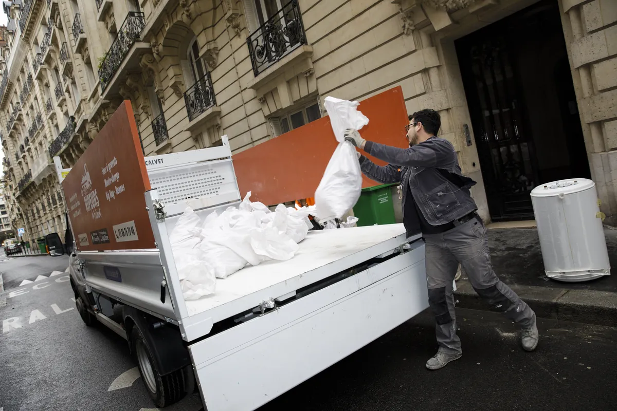 Ripeur chargeant un sac à gravats sur le plateau d'un camion Les Ripeurs dans une rue haussmannienne, ballon d'eau chaude posé au sol