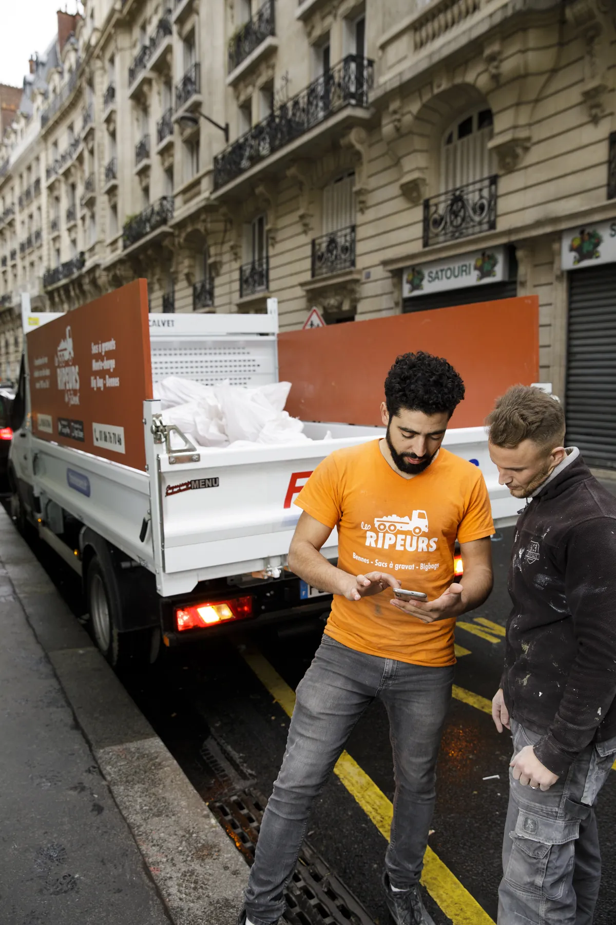 Ripeur et artisan client consultant ensemble un smartphone devant un camion chargé de sacs, dans une rue