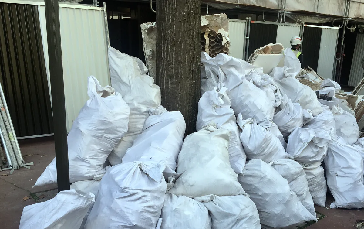 Sacs à gravats blancs empilés au pied d'un arbre devant une palissade de chantier