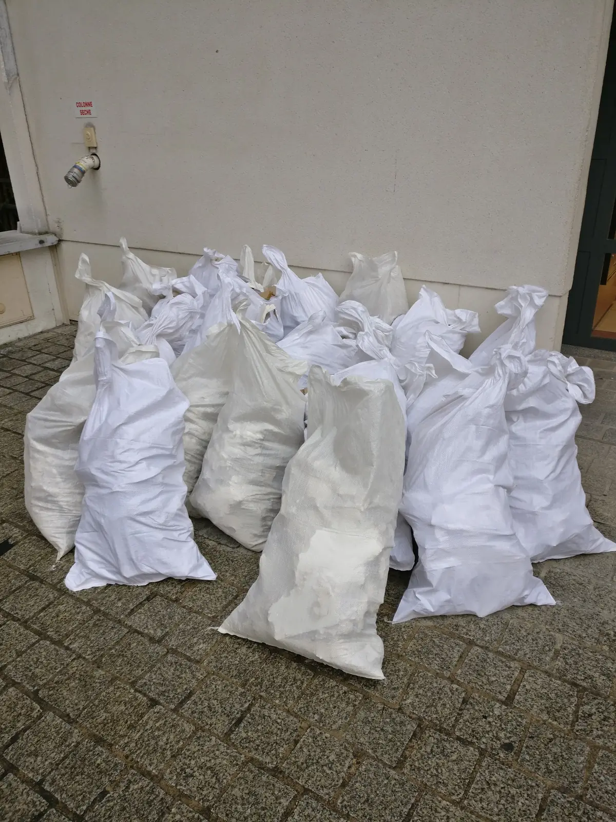 Sacs blancs de gravats regroupés au pied d'un immeuble sur un sol pavé