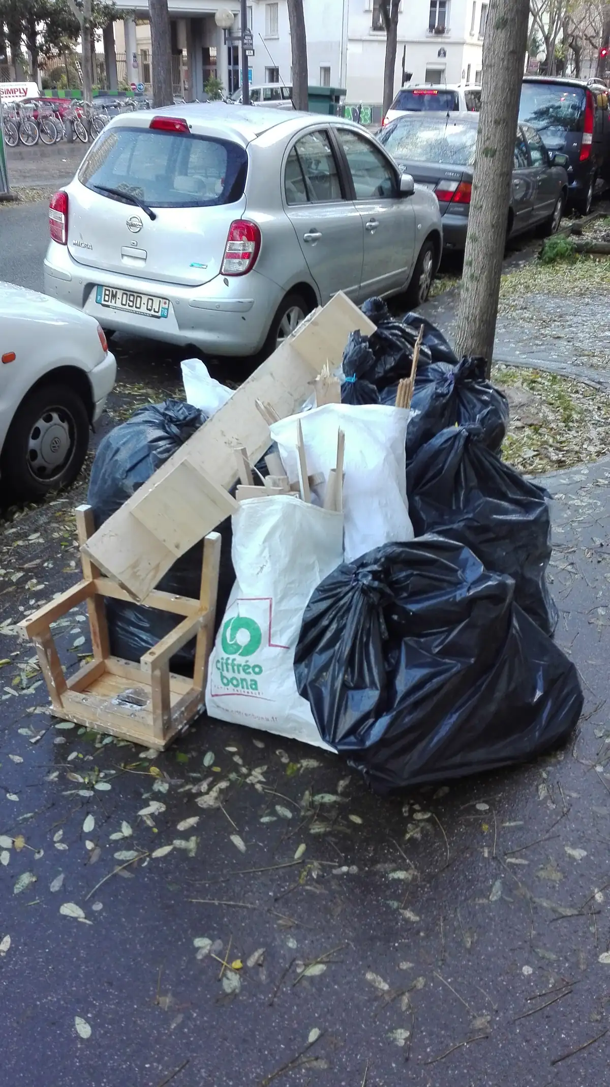 Sacs noirs et sac blanc, tréteau en bois et chutes de tasseaux déposés sur un trottoir le long de voitures garées
